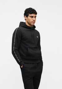 Άνδρας με σγουρά μαλλιά φορούντας μαύρη φούτερ με κουκούλα και λογότυπο, και ταιριαστό μαύρο παντελόνι, με τα χέρια στις τσέπες, πάνω από ένα απλό λευκό φόντο.