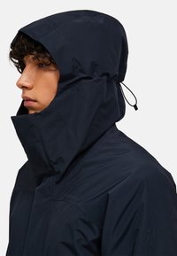 Marine waterproof jack met een hoge kraag en verstelbare capuchon. Het heeft een gladde textuur en een minimalistisch ontwerp, dat functionaliteit benadrukt.