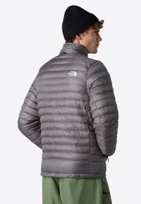 Šedá zateplená péřová bunda s vysokým límcem, horizontálním prošíváním a malým bílým logem na zadní straně. Nositá přes zelené kalhoty.