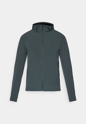 Mørk grå Nike-jakke med full glidelås, høy krage og hette. Har lange ermer og to frontlommer. Glatt, lett stoff.