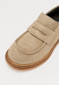 Zapato de ante beige con una correa ancha, punta redondeada y una superficie texturizada. Presenta costuras de color claro y una suela de madera.