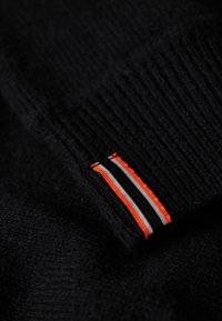 Dettaglio di un maglione nero a maglia con polsino a costine e un'etichetta a strisce multicolori in arancione, nero e bianco. Texture morbida, design casual.