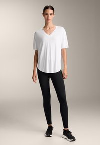 Bílé tričko s krátkým rukávem a V výstřihem, se zaobleným lemem, v kombinaci s černými legínami a černými sportovními botami, zasazené na neutrálním pozadí.