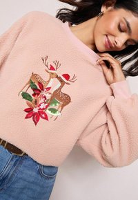 Felpa in pile rosa con scollo rotondo, decorata con cervi ricamati che indossano cappelli di Babbo Natale e fiori di poinsettia sulla parte anteriore.