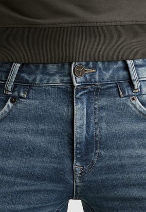 Close-up van blauwe spijkerbroek met vastgeknoopte knoop en donkerribgebreide tailleband van een kledingstuk erboven.