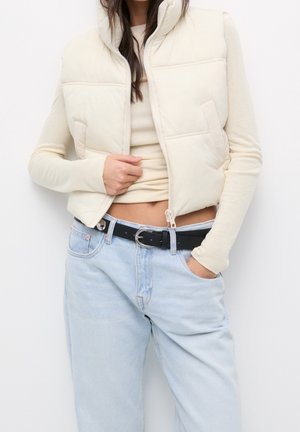 Kvinde iført en cremefarvet polstret vest over en langærmet cremefarvet top, lyseblå jeans med sort bælte og hånden i lommen.
