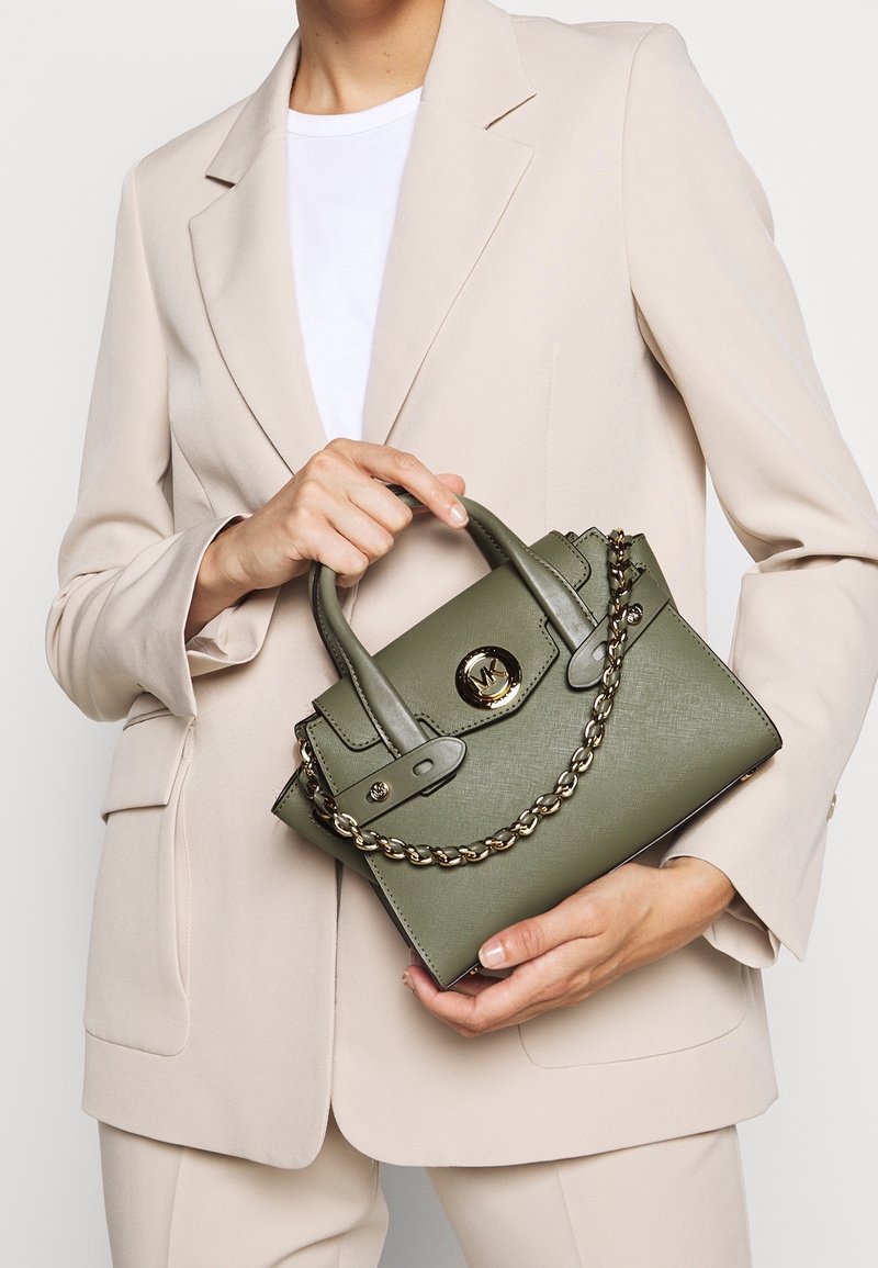 Personne en costume beige tenant un sac à main vert olive avec une chaîne dorée et une boucle métallique ronde avec logo.