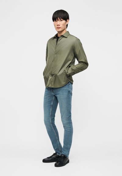 Jeune homme debout avec les mains dans les poches, portant une veste boutonnée vert olive, un jean bleu clair et des baskets noires sur un fond gris uni.