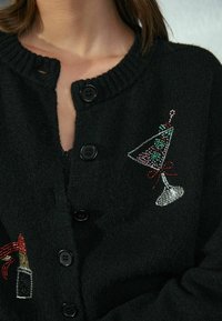 Cardigan negru cu guler ribstit, prezentând designuri brodate de cocktailuri și artificii din mărgele colorate. Cinci nasturi negri în față.