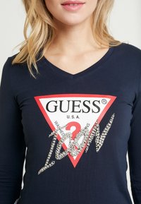 Žena v námořnické modré košili s V-výstřihem, na které je červené a bílé logo Guess a lesklý text s kamínky, který říká "Ikona."