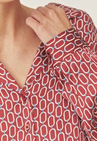 Bluză roșie cu model format din lanțuri albe și albastre, cu guler și detalii cu nasturi, realizată dintr-un material ușor.