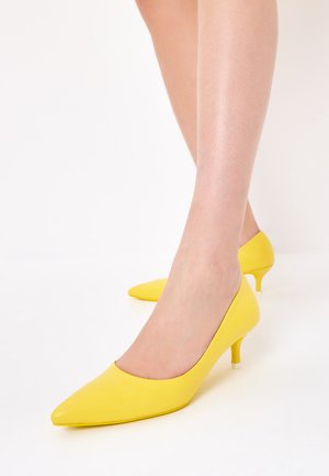 Scarpe alti tacchi in pelle gialla con punta appuntita, tacco basso e superficie liscia, indossate su una gamba scoperta.