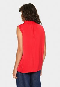 Top rosso senza maniche con colletto alto, texture liscia e dettagli di piega sul retro, abbinato a pantaloni larghi in denim blu.