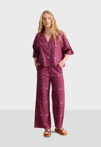 Tweedelig outfit met een kortebloes met knopen en wijde broek, beide in een paarse ikatprint met een glanzende textuur.