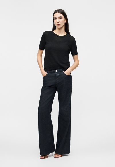 Γυναίκα με μακριά σκούρα μαλλιά, που φοράει χρυσούς κρίκους στα αυτιά, μαύρο μπλουζάκι με κοντά μανίκια, σκουρόχρωμα τζιν με φαρδιά παντελόνια και πέδιλα με ανοιχτά δάχτυλα, με τα χέρια στις τσέπες.