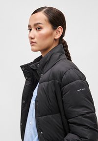 Črna napihljiva jakna z visokim ovratnikom, z zadrgo in pritisnimi gumbi. Teksturnana površina s prešito vzorci in logotipom na rokavu.