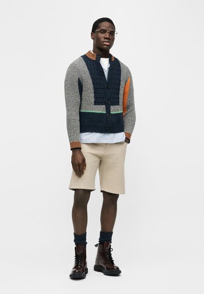 Άνδρας με γυαλιά, πολύχρωμη πλεκτή ζακέτα, ανοιχτόχρωμα σορτς, σκούρα μποτάκια με σούρες στις κάλτσες, στέκεται μπροστά σε απλό φόντο.