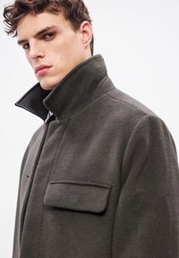Σκούρο γκρι μάλλινο παλτό με ψηλό γιακά, που διαθέτει μία μονή τσέπη στο στήθος και λεία υφή, σχεδιασμένο για εφαρμοστή σιλουέτα.