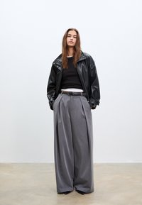 Crna kožna jakna s pritisnim ovratnikom, crni pulover, pružene hlače sive boje visoka struk, i crne cipele u minimalističkom okruženju.