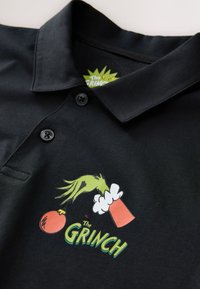 Musta poolopaita, jossa on värikäs grafiikka, jossa Grinchin käsi pitää koriste-esinettä, ja alla vihreällä tekstillä "The Grinch".