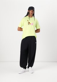 Neon žluté tričko s barevným grafikou, v kombinaci s volnými černými kalhotami a bílou sportovní obuví. Model má na sobě černou čepici.
