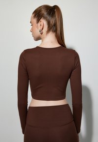 Brązowy krótki top z długim rękawem, wykonany z gładkiego materiału, z okrągłym dekoltem i dopasowanym krojem. W połączeniu ze spodniami w kolorze brązowym o wysokim stanie.