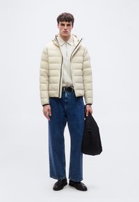 Άνδρας που φοράει μπεζ μπουφάν, ανοιχτό μπεζ πουκάμισο με φερμουάρ, φαρδιά μπλε τζιν παντελόνια, μαύρη ζώνη και παπούτσια, κρατώντας μια μαύρη τσάντα μπροστά σε απλό φόντο.