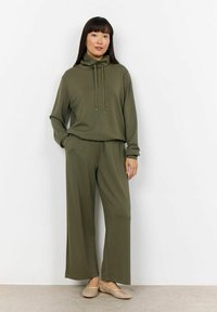 Felpa verde oliva a maniche lunghe con lacci abbinata a pantaloni a gamba larga. Tessuto morbido, vestibilità rilassata e accentuata da calzature leggere.
