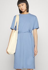 Vestido de t-shirt de algodão azul claro com mangas curtas, detalhe de cintura com nó e fenda lateral, acompanhado de uma mala tote bege.