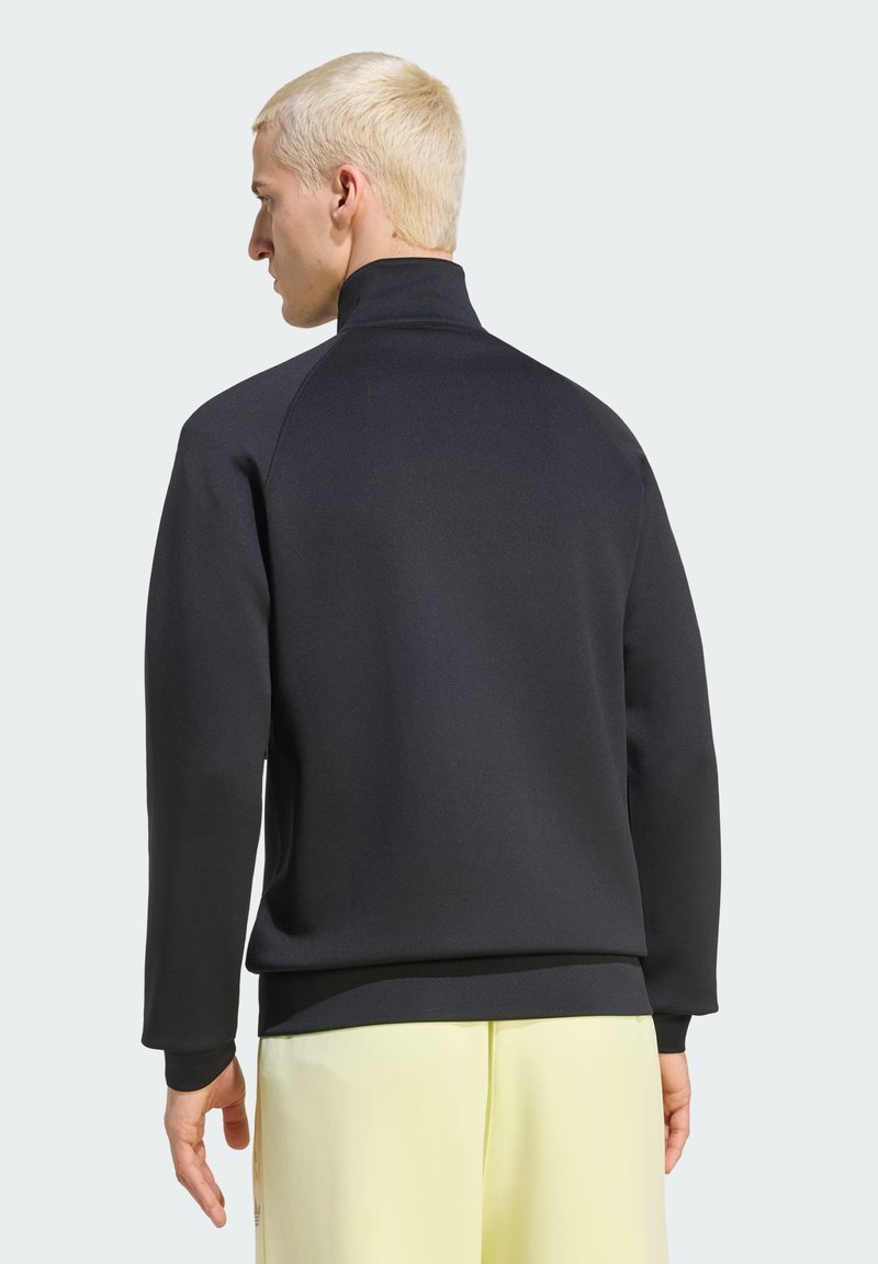 Mann mit kurzem blonden Haar, der eine schwarze Langarmjacke und blassgelbe Hose trägt, von hinten vor einfarbigem Hintergrund gezeigt.