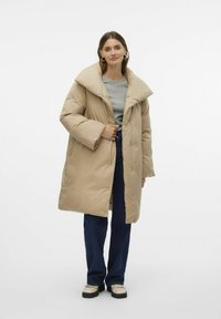 Beige pufferjakke med stor krave, trykknapper og quiltet tekstur. Båret over en grå top og parret med mørke jeans og lyse sko.