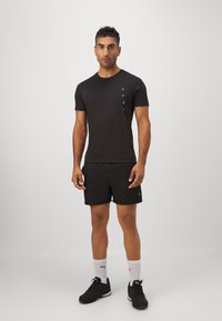 Camisola atlética preta com um pequeno logótipo branco no lado esquerdo do peito, combinada com calções pretos e sapatos pretos. Design simples e ajustado.