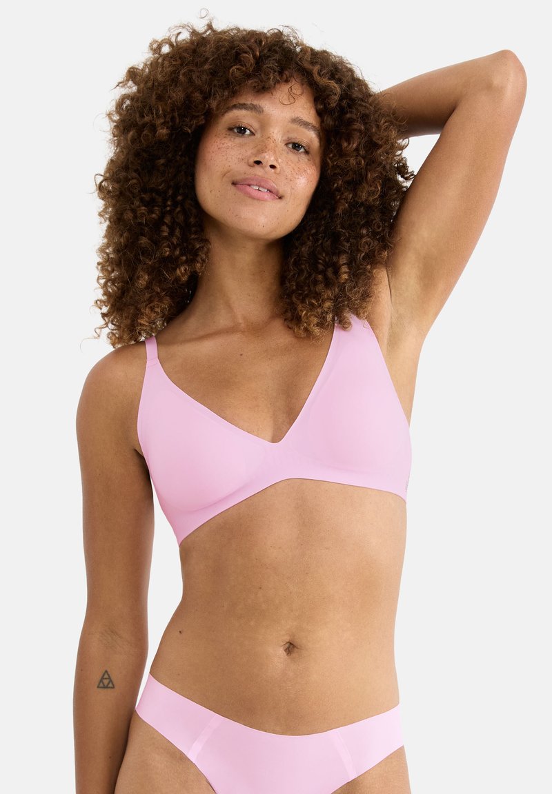 Femme aux cheveux bouclés portant un soutien-gorge sans coutures rose clair et un sous-vêtement assorti, posant avec un bras levé sur un fond blanc.