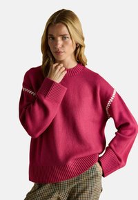 Pulover tricotat fucsia cu manșete și borduri cu nervuri, având accente decorative albe pe mâneci, purtat peste pantaloni în carouri.