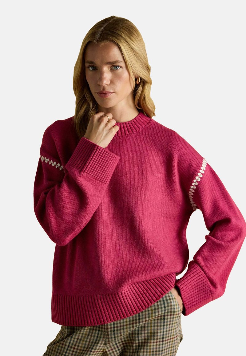 Pulover tricotat fucsia cu manșete și borduri cu nervuri, având accente decorative albe pe mâneci, purtat peste pantaloni în carouri.