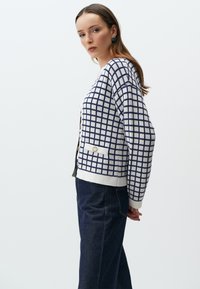 Gehaakte cardigan in ivoorkleur met een marineblauw ruitpatroon, voorzien van een v-hals, knopen aan de voorkant en zijzakken. Gepaard met donkerblauwe denim.