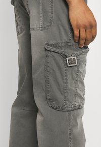 Graue Cargohosen mit strukturiertem Stoff, seitlicher Tasche mit Klappe und silberner Schnalle sowie sichtbaren Nähten entlang der Nähte.