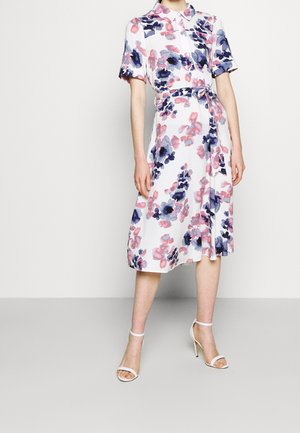 Kobieta ubrana w białą sukienkę midi z różowo-fioletowym kwiatowym wzorem, z krótkimi rękawami, kołnierzykiem, paskiem i białe sandały na wysokim obcasie z paskiem wokół kostki.