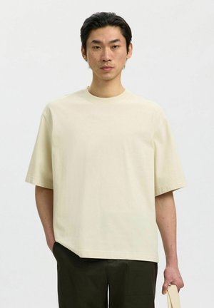 Jeune homme aux cheveux noirs porte un t-shirt beige ample et un pantalon foncé, tenant un objet beige dans sa main droite sur un fond uni.