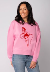 Un sweat-shirt rose à coupe ample, présentant un graphique rouge d'une femme avec un lasso. Poignets et ourlet côtelés, associé à un jean bleu.
