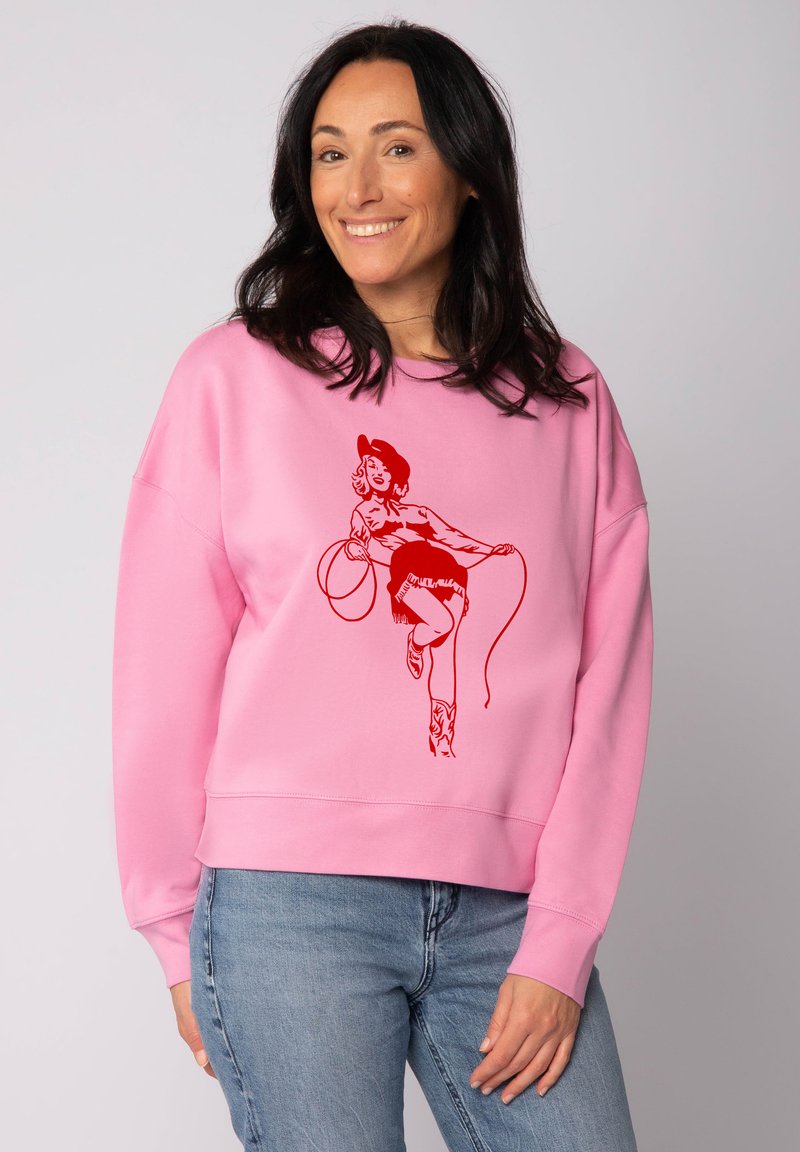 Un sweat-shirt rose à coupe ample, présentant un graphique rouge d'une femme avec un lasso. Poignets et ourlet côtelés, associé à un jean bleu.