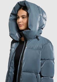 Minkšta striukė blizgiu mėlynos skalūno spalvos atspalviu su dideliu, struktūruotu gobtuvu. Yra užtrauktukas priekyje ir šoninės kišenės, išryškinančios siūtas detalės tekstūrą.