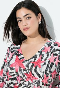 Jonge vrouw met donker haar die zilveren oorbellen draagt en een V-hals top met roze zeesterren en zwart-wit zebra patronen.