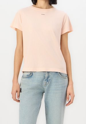 Femme portant un t-shirt à manches courtes rose clair avec le texte "PINKO" près du col et un jean en denim bleu clair sur fond blanc.