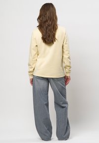 Persoon met lang bruin haar, gekleed in een crèmekleurige trui en loszittende, vervaagde grijze jeans, staand met de rug naar de camera tegen een effen achtergrond.