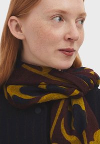 Knitted scarf featuring a maroon and yellow abstract pattern on a dark background. Soft texture with a loose, draping shape.