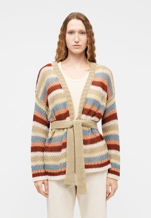 Mujer con un cárdigan de punto multicolor a rayas con cinturón en beige, óxido, azul y crema, sobre una camisa blanca y pantalones claros.