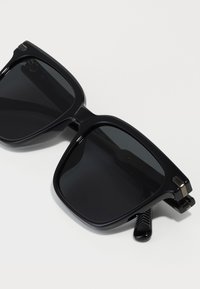 Black sunglasses with a square design, dark lenses, and a sleek plastic frame. Features metallic accents on the temples and textured hinges.