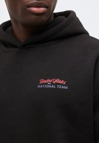 Gros plan d'une personne portant un sweat à capuche noir avec l'inscription "United States Men's National Team" brodée sur la poitrine en rouge et bleu.