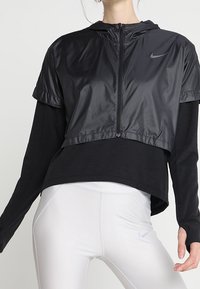 Veste noire courte en tissu léger et brillant avec capuche ; portée sur un haut noir ajusté à manches longues. Leggings blancs avec détail logo.